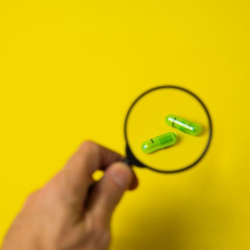 From above of crop unrecognizable male holding magnifying glass over green pills on yellow background in studio — Biggest Boo