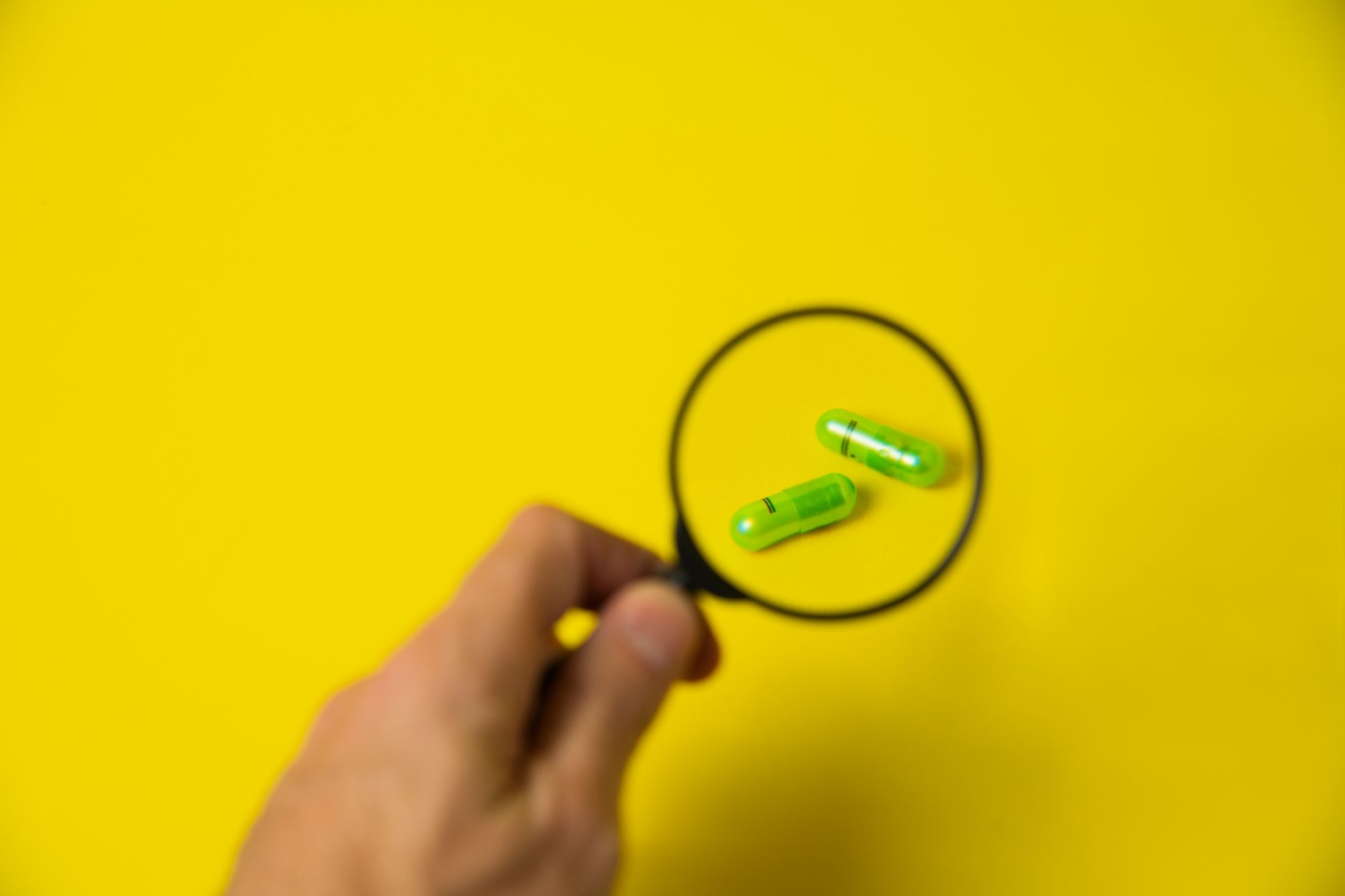 From above of crop unrecognizable male holding magnifying glass over green pills on yellow background in studio — Biggest Boo