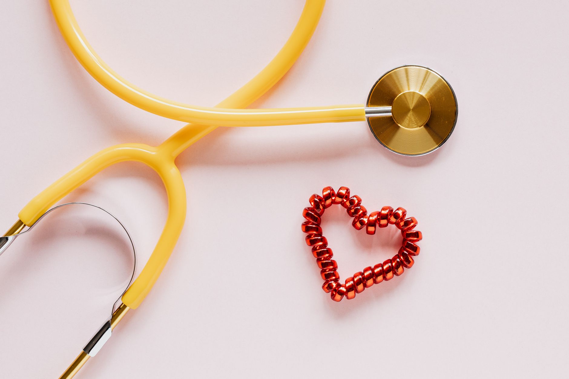 Top view of yellow stethoscope made of plastic and metal material near small handmade heart of spiral hair tie on pale pink b