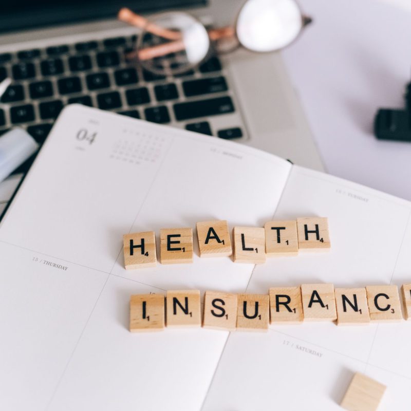 Close-up of Scrabble tiles spelling 'Health Insurance' on a planner with a laptop. — I'm a Girl and My Hair is Thinning withi