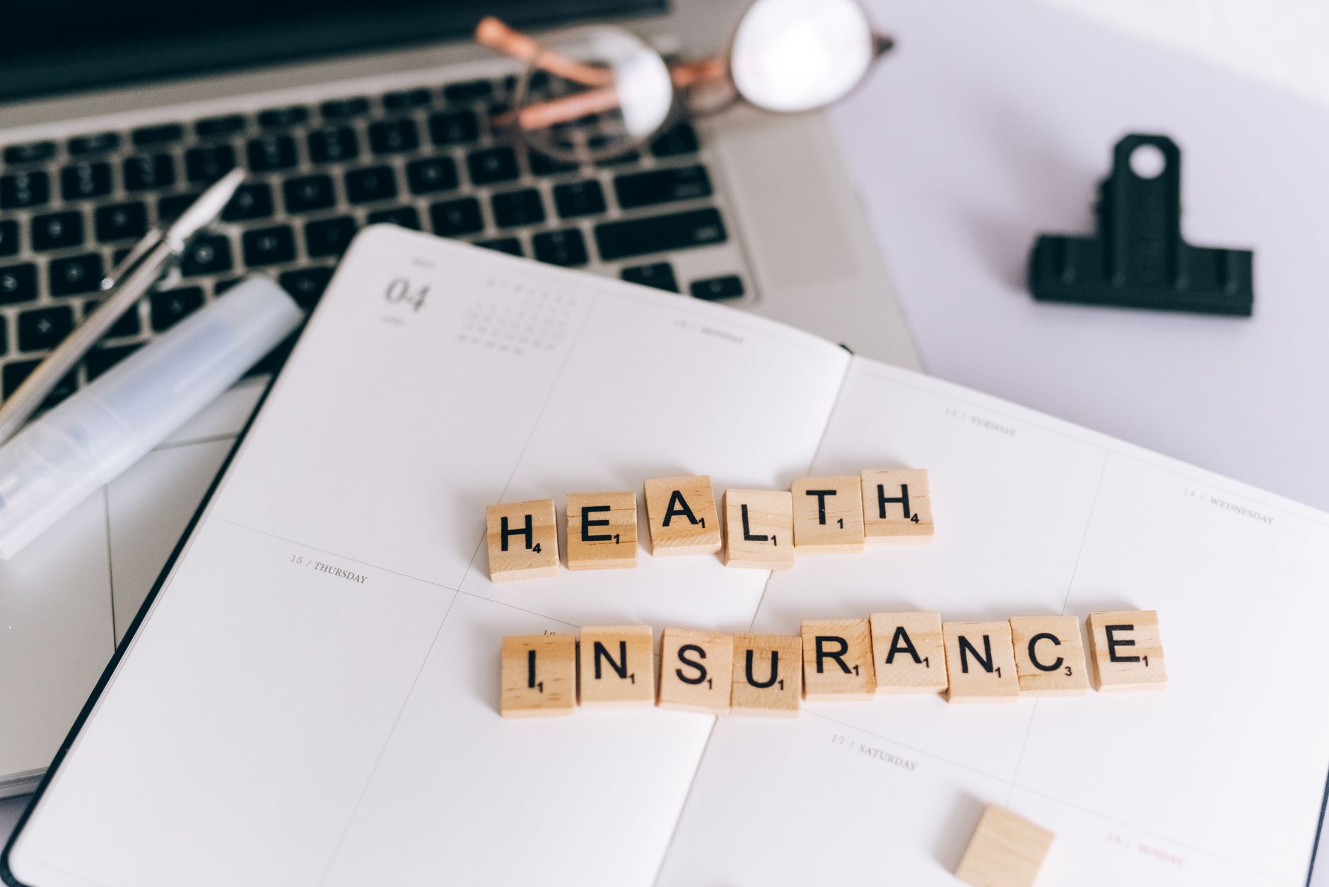 Close-up of Scrabble tiles spelling 'Health Insurance' on a planner with a laptop. — I'm a Girl and My Hair is Thinning withi