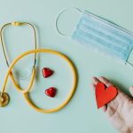 Top view of crop anonymous person hand with red paper heart on table with stethoscope and medical mask for coronavirus preven